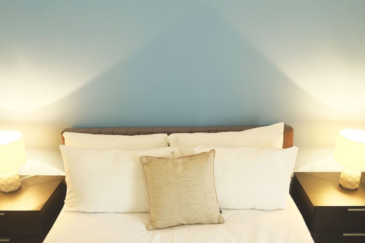 Cama bem arrumada com roupa de cama branca e almofada bege, com duas mesas de cabeceira com abajures, em frente a uma parede azul.