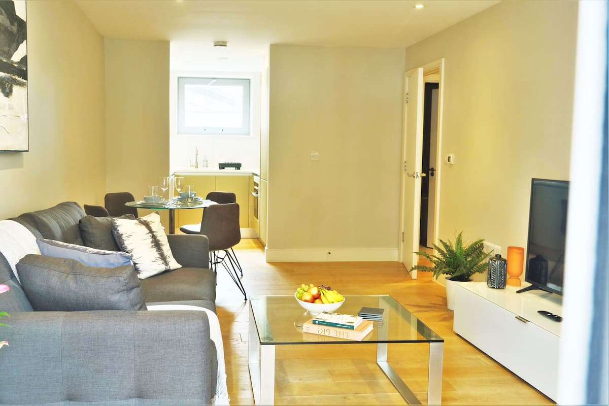 Uma sala de estar moderna com um sofá cinzento, uma mesa de centro de vidro e uma área de refeições ao fundo.