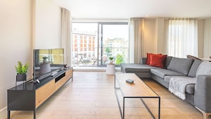 A modern living room with a flat-screen TV, a grey sofa, a coffee table, and a view of the city through large windows. - Accomodo London Southbank - Spacious & Elegant Apartments near Big Ben & London Eye (London)