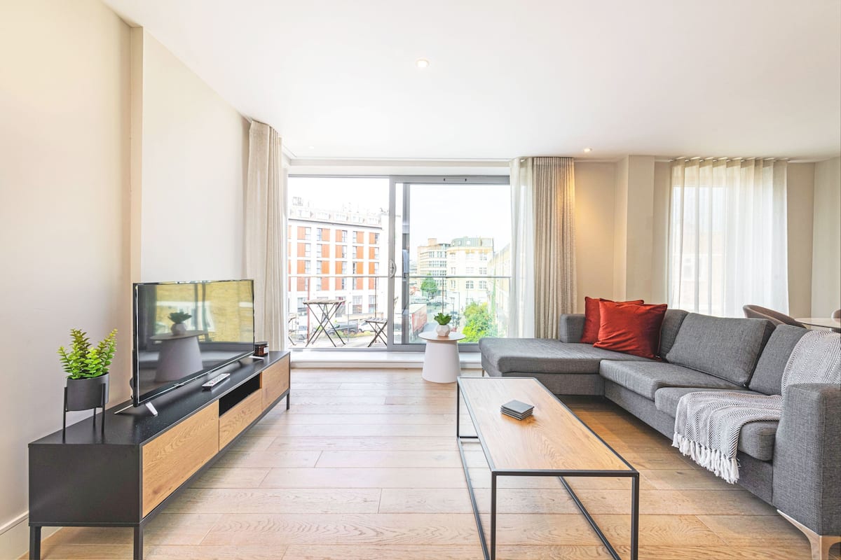 Uma sala de estar moderna com televisão de tela plana, um sofá cinza, uma mesa de centro e vista da cidade por grandes janelas.