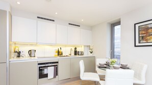 A modern kitchen with white cabinetry, a built-in oven, and a dining area with white chairs and a glass table. - Accomodo London Southbank - Spacious & Elegant Apartments near Big Ben & London Eye (London)