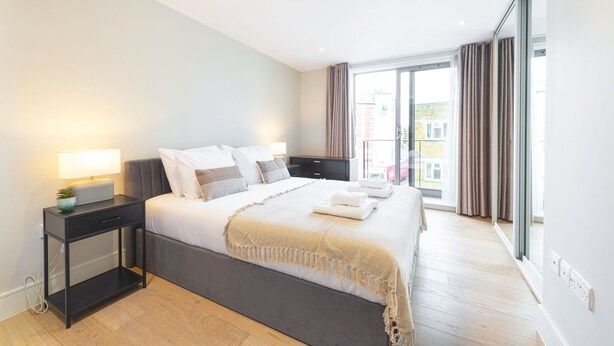 A modern bedroom with a large bed, bedside tables, a lamp, and a view of the city through large windows. - Accomodo London Southbank - Spacious & Elegant Apartments near Big Ben & London Eye (London)