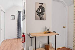 A hallway with a wooden floor, a fire extinguisher, a wall-mounted shelf with kitchen utensils, and a framed artwork.