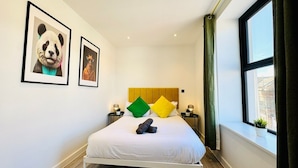 A neatly made bed with a white duvet, green and yellow pillows, and a rolled-up towel. There are two framed pictures on the wall, a window with a view of buildings, and a small plant on the windowsill. - Central Bournemouth | 2-Bed, 4 Guests | Prime Spot (Bournemouth, Christchurch and Poole)