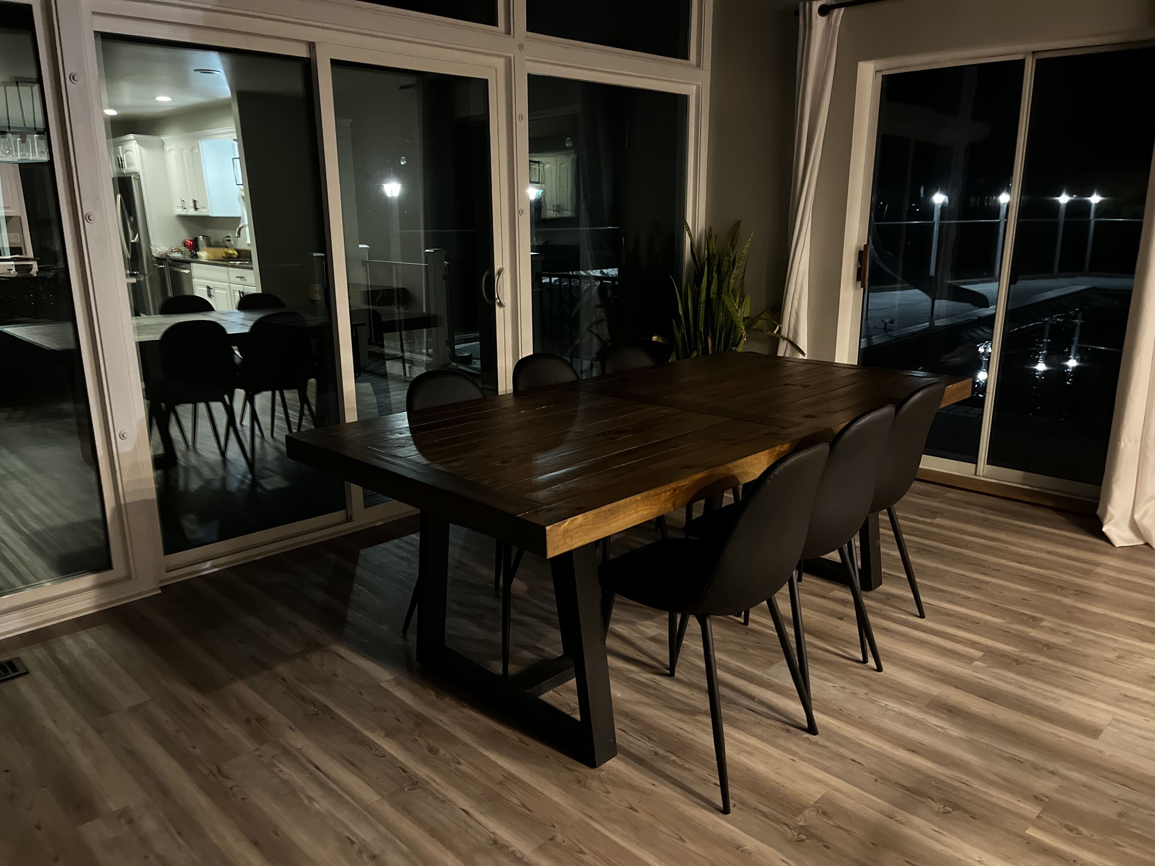 Un comedor con mesa y sillas de madera, un divisor corredizo de vidrio que da a una cocina, y vista nocturna de la ciudad.