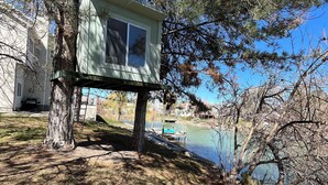 Une cabane dans les arbres avec une fenêtre, entourée d’arbres et bordée par un lac.