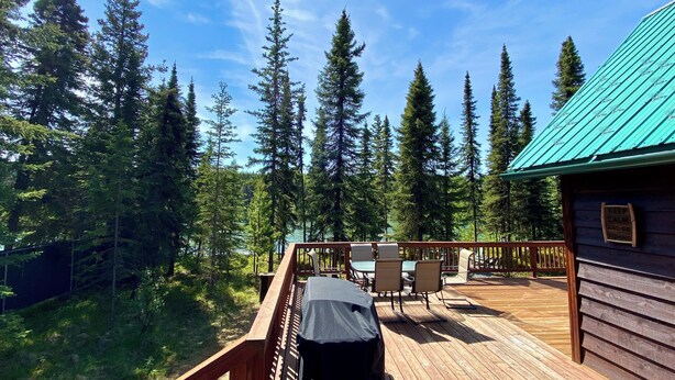 A wooden deck with outdoor seating and a view of tall evergreen trees. - Kenai Riverfront Cabin! Fish, Explore, & Relax! (Sterling)
