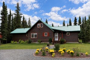 A wooden cabin with a green roof, surrounded by a well-maintained lawn and a gravel driveway. - Kenai Riverfront Cabin! Fish, Explore, & Relax! (Sterling)
