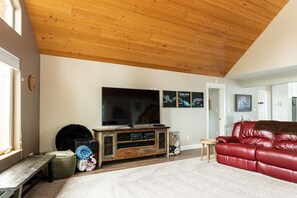 A living room with a wooden ceiling, a flat-screen TV, a red leather sofa, and a small round table. - Kenai Riverfront Cabin! Fish, Explore, & Relax! (Sterling)