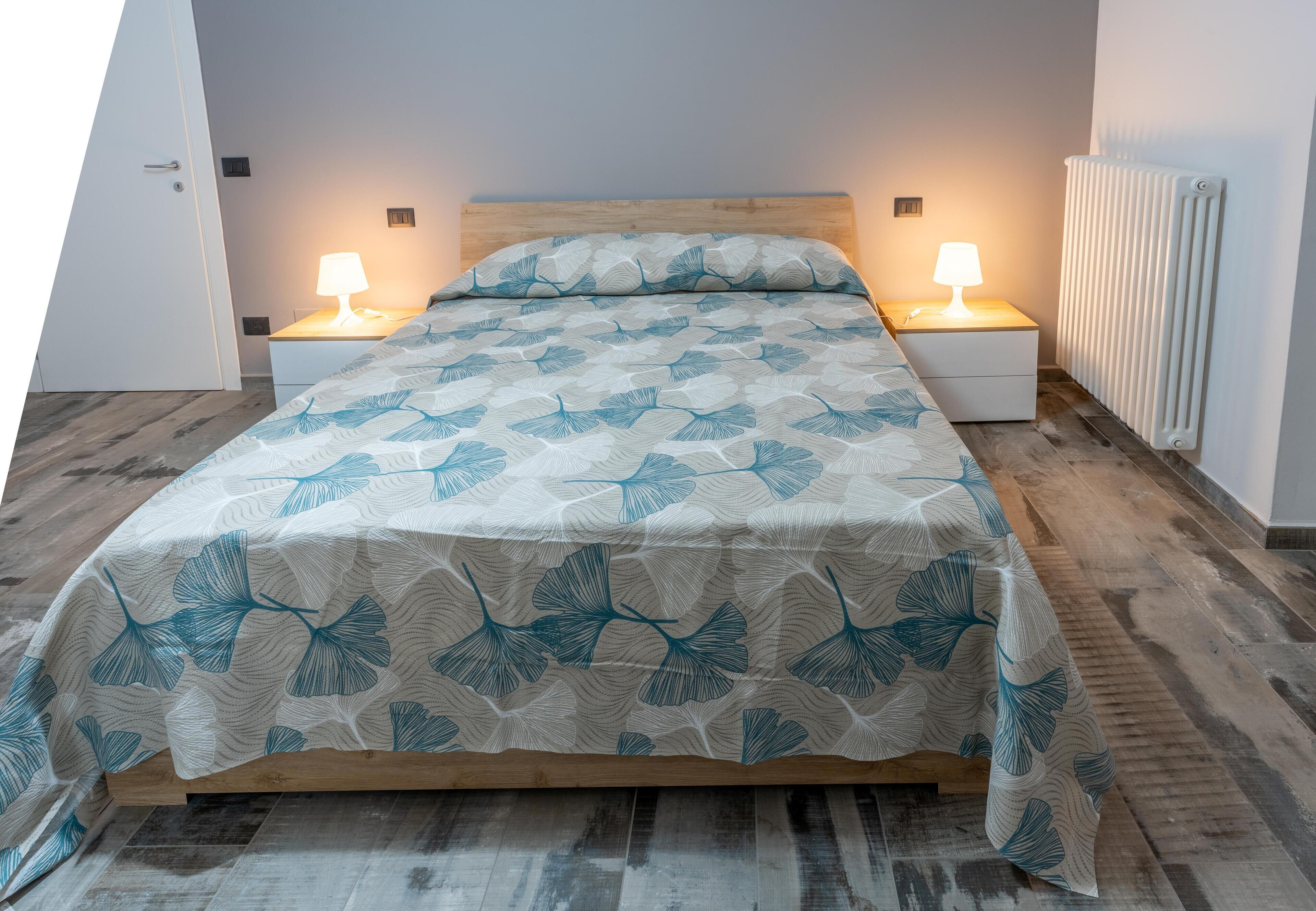 A bed with a leaf-patterned bedspread, two bedside lamps, a wooden headboard, and a radiator on the wall.
