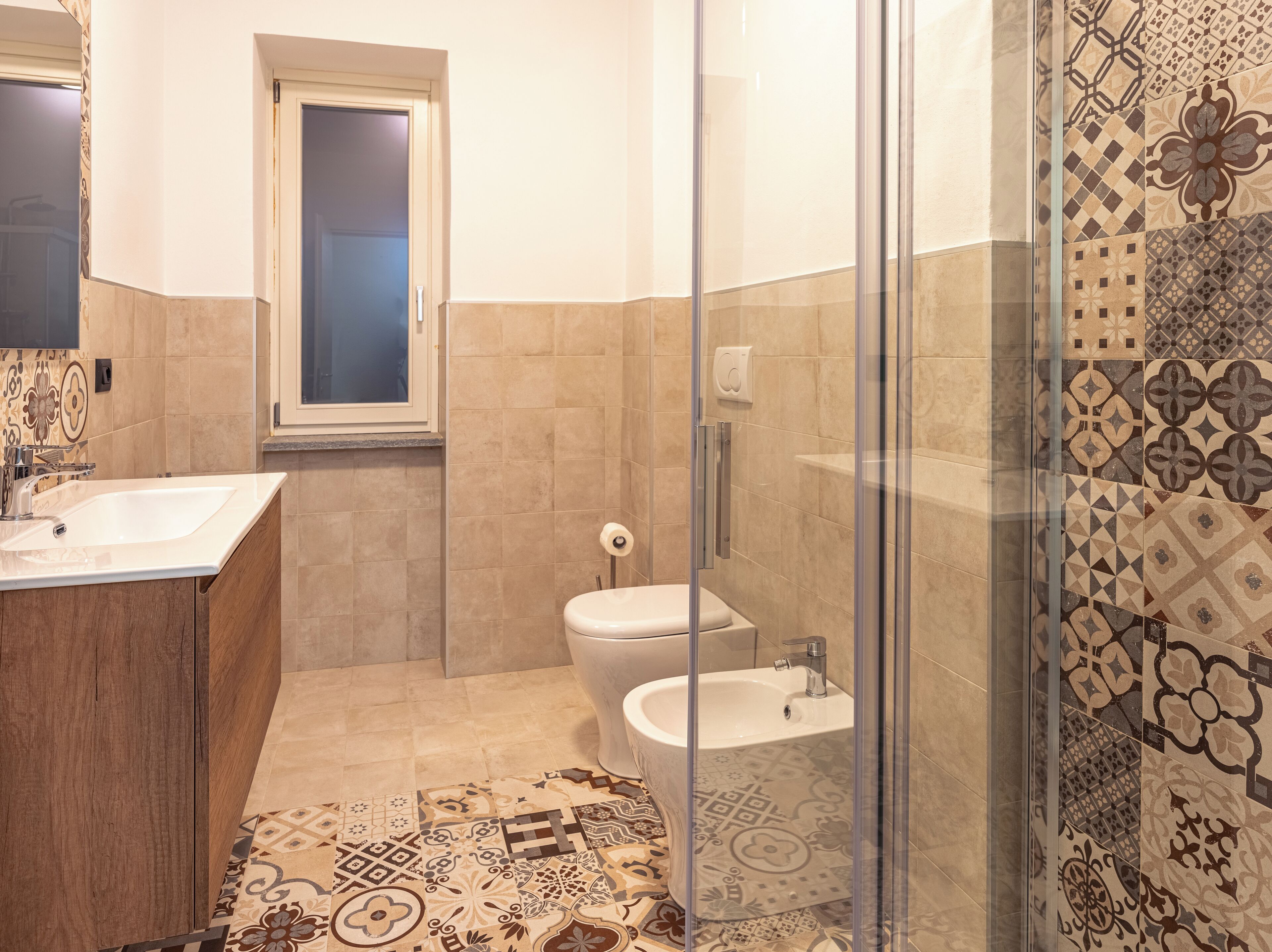 Casa de banho com chuveiro, sanita e lavatório, com azulejos estampados e armário de madeira.