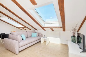Un dormitorio tipo loft moderno con techo inclinado, tragaluces, vigas de madera, un sofá, una mesita blanca, un televisor y dos jarrones verdes.
