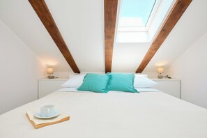Une chambre avec une lucarne, un lit recouvert de draps blancs, deux tables de chevet avec des lampes, et une tasse et une soucoupe posées sur le lit.