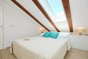 Un dormitorio con techo inclinado, tragaluz, cama con edredón blanco y azul turquesa, y lámparas de noche.
