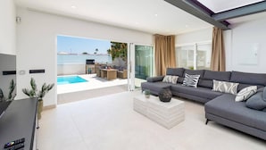 En moderne stue med en grå sofa, et sofabord og et fladskærms-tv. Der er en skydedør af glas, der fører til et udendørs område med en pool og siddepladser.