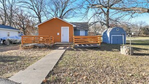 A small, single-story building with a front porch and a door, surrounded by a wooden fence and a concrete path leading to it. - Clean, quiet 2BR home with utilities included and fast WiFi. Comfortable setup with open surroundings and easy access to downtown Kansas City (about 15 minutes). Ideal for short stays, work trips, or a simple, reliable place to unwind. (Sugar Creek)