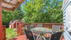 A covered outdoor deck with a glass table and chairs, surrounded by a red railing and a small shed in the background.