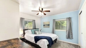 A bedroom with a bed, a desk, a ceiling fan, and two windows with curtains.