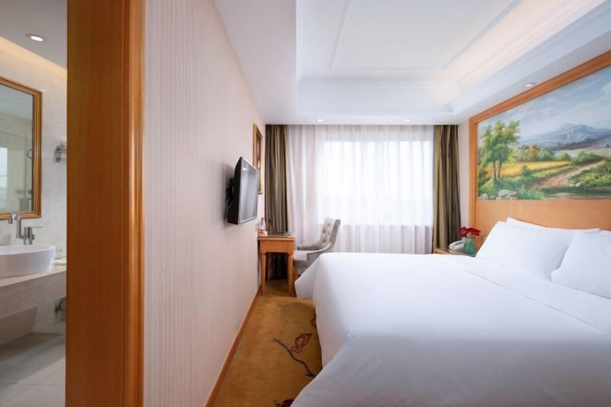 Habitación de hotel con una cama grande, televisor montado en la pared, escritorio con silla, un cuadro en la pared y baño con lavabo y espejo.