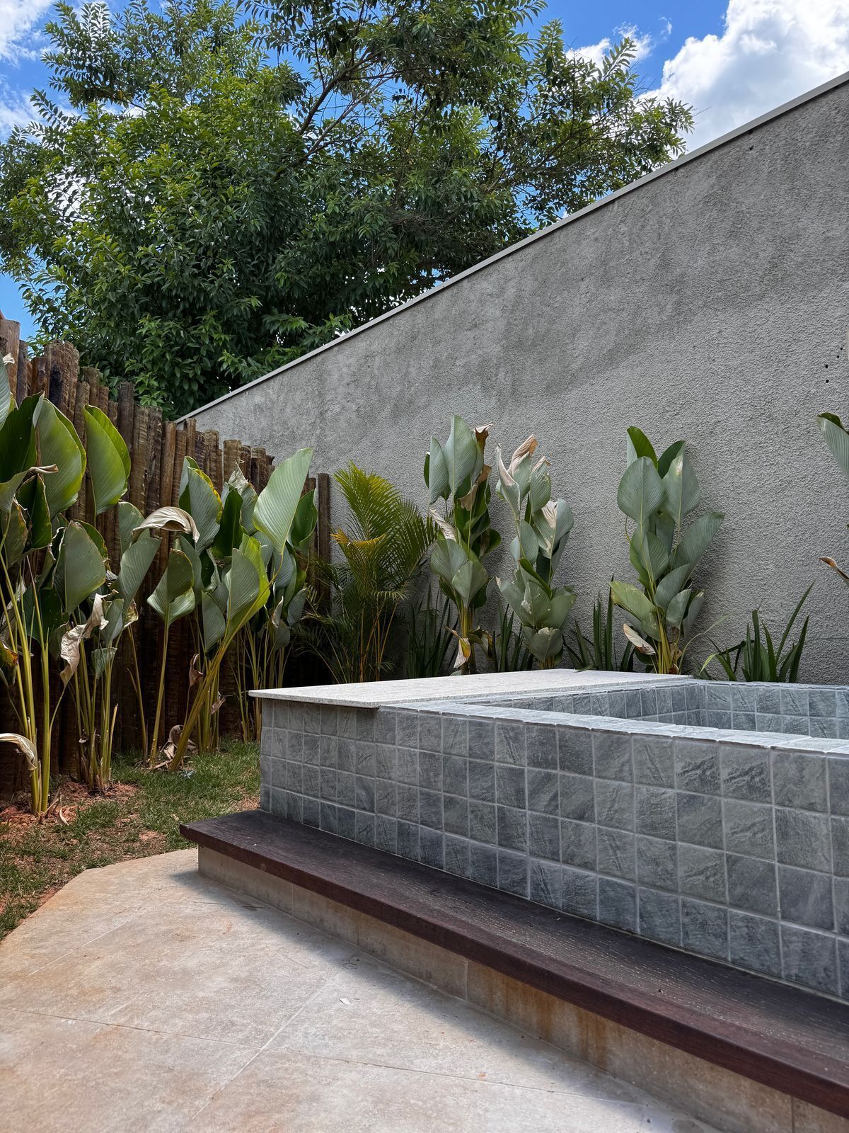 Zona exterior moderna amb un mur de formigó, una banca enrajolada i diverses plantes tropicals.