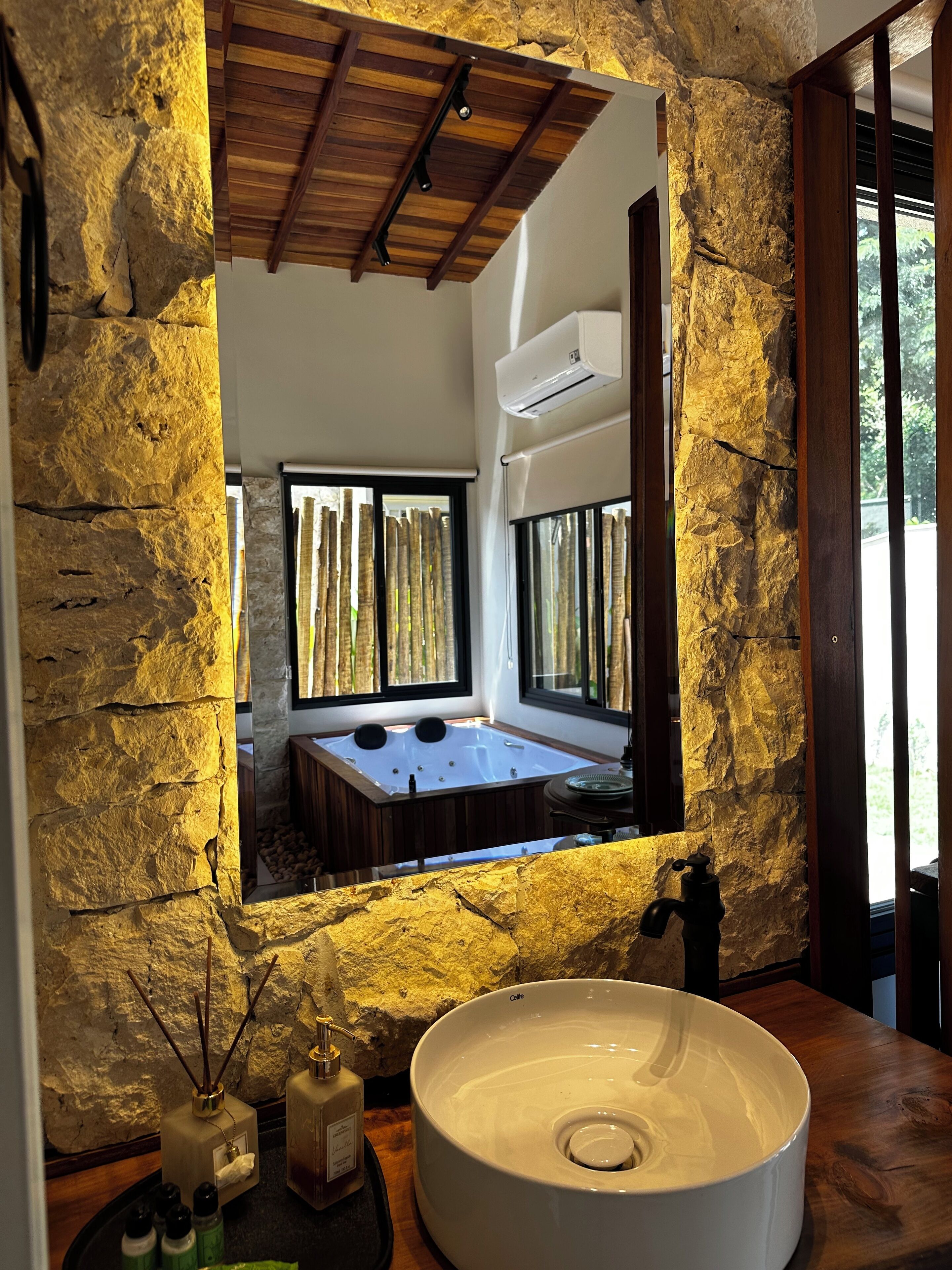 A bathroom with a stone wall, a large sink, a bathtub, and a window with vertical blinds.