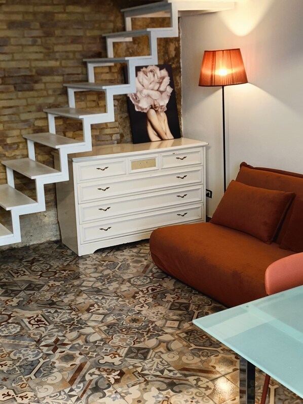 A room with a patterned floor, a white dresser with drawers, a brown sofa, a glass table, and a staircase with a picture frame. - Historic Apartment (Agrigento)