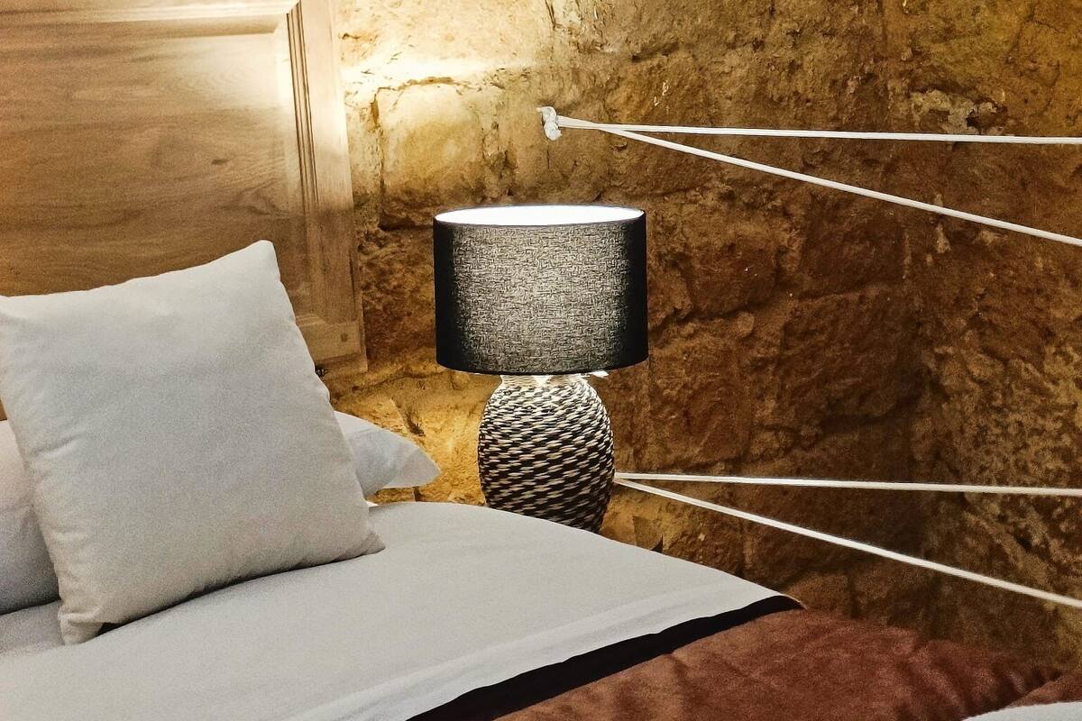 Ein Schlafzimmer mit rustikaler Steinwand, einem Holzkopfteil, einem Bett mit weißer und brauner Bettwäsche und einer Lampe mit geflochtenem Fuß.