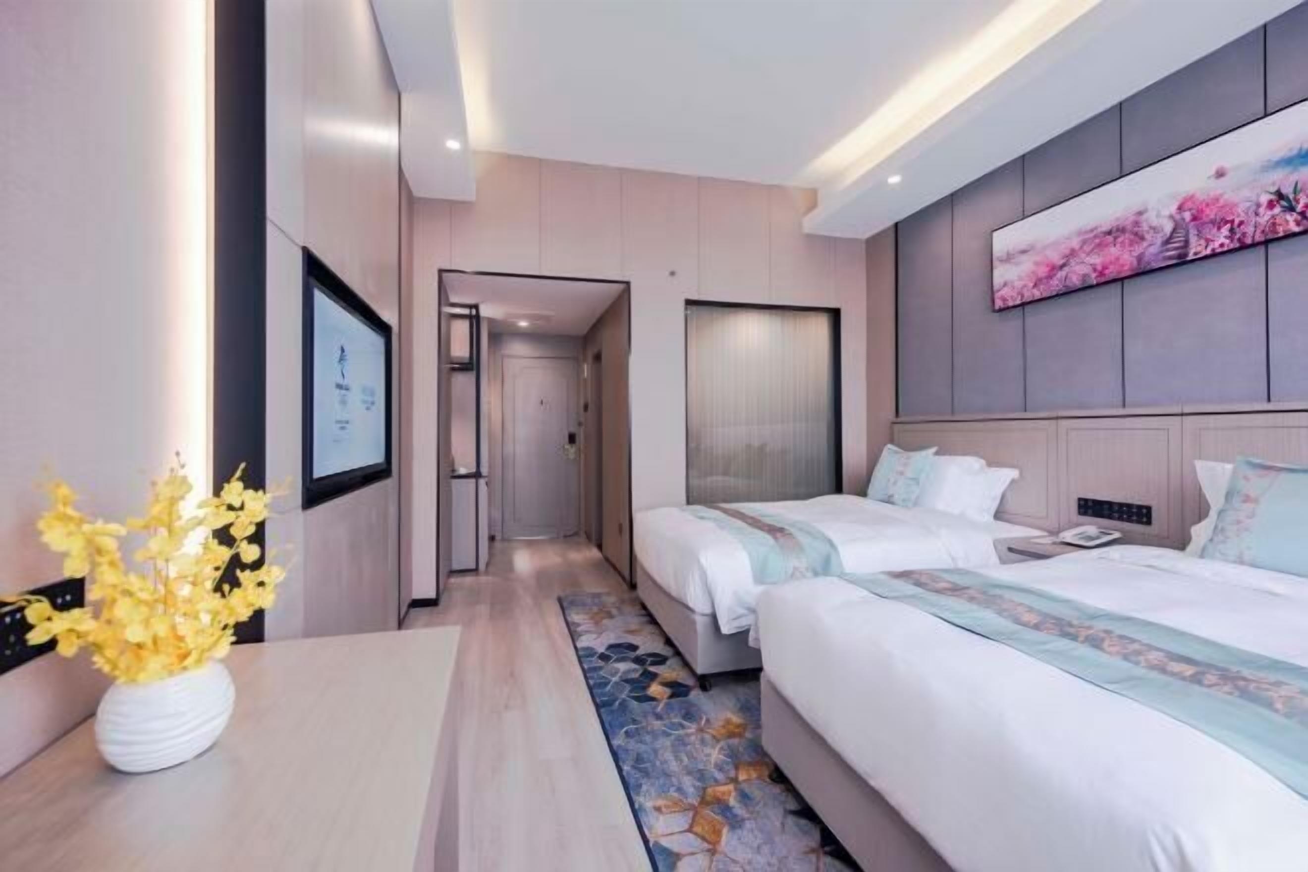 A hotel room with two beds, a bedside table with a vase of flowers, a large painting on the wall, and a door leading to another room.