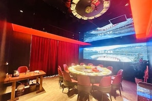 A conference room with a large screen displaying a stadium, a round table with chairs, and a red curtain backdrop. - Qianmei Hotel (Ganzhou)