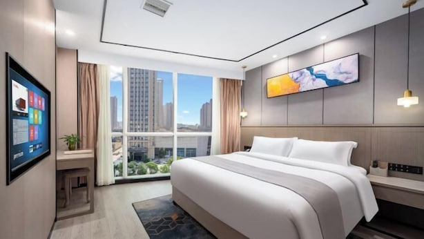 Een moderne hotelkamer met een groot bed, een bureau met een computer, een televisie en een groot raam met uitzicht op de stad.