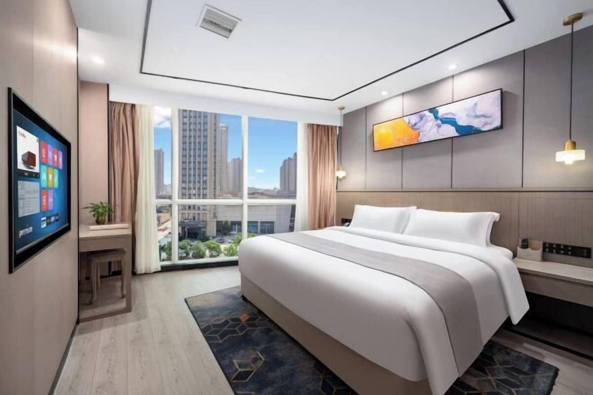 A modern hotel room with a large bed, a desk with a computer, a TV, and a large window with a city view.
