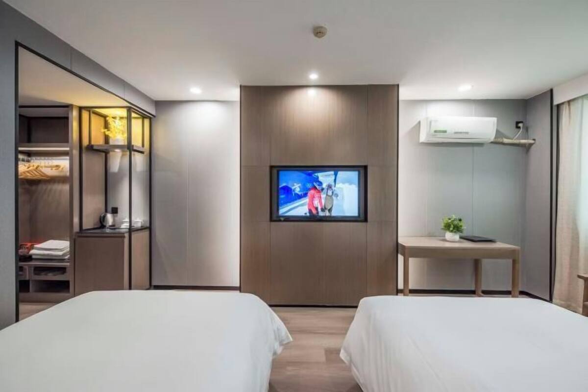 A modern hotel room with two beds, a TV mounted on the wall, a small table, and a wardrobe with a mirror.