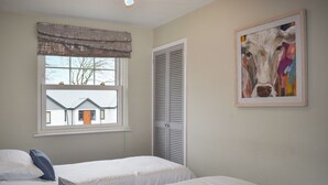 Un dormitorio con dos camas, una ventana con vista a una casa, una puerta con contraventanas y un cuadro en la pared.