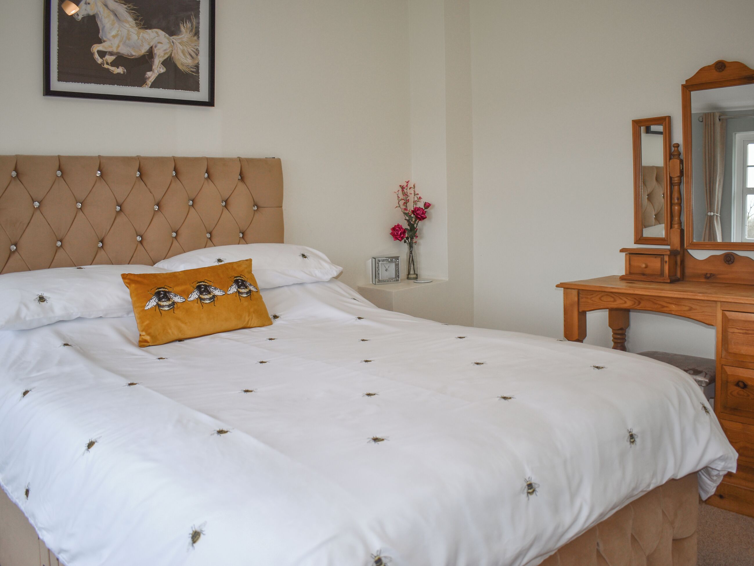 Uma cama bem arrumada com cabeceira, mesa de cabeceira com um vaso de flores e cômoda de madeira com espelho.