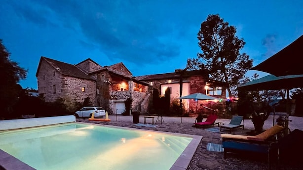 A swimming pool with lounge chairs and umbrellas, surrounded by stone buildings and a large tree.
