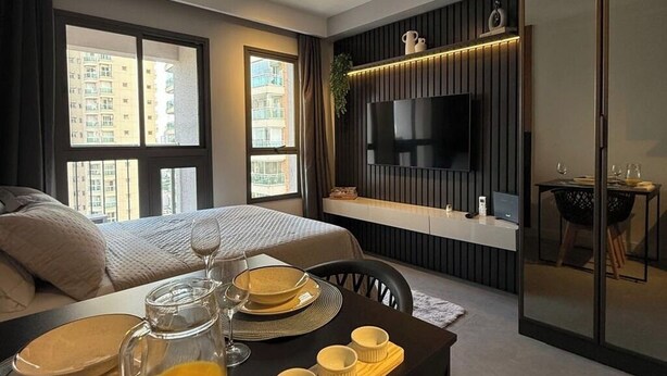 Una habitación de hotel moderna con una cama, una mesa de comedor con platos y vasos, un televisor montado en la pared y ventanas grandes con vistas a la ciudad.