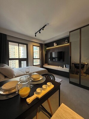 Una moderna camera d'albergo con un letto, un tavolo da pranzo apparecchiato con piatti e bicchieri, una TV a parete e ampie finestre con vista sulla città.