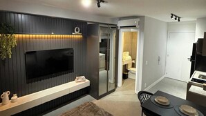 Un dormitorio moderno con una cama grande, un televisor montado en una pared oscura, una mesita con platos, y una puerta que da a un baño.