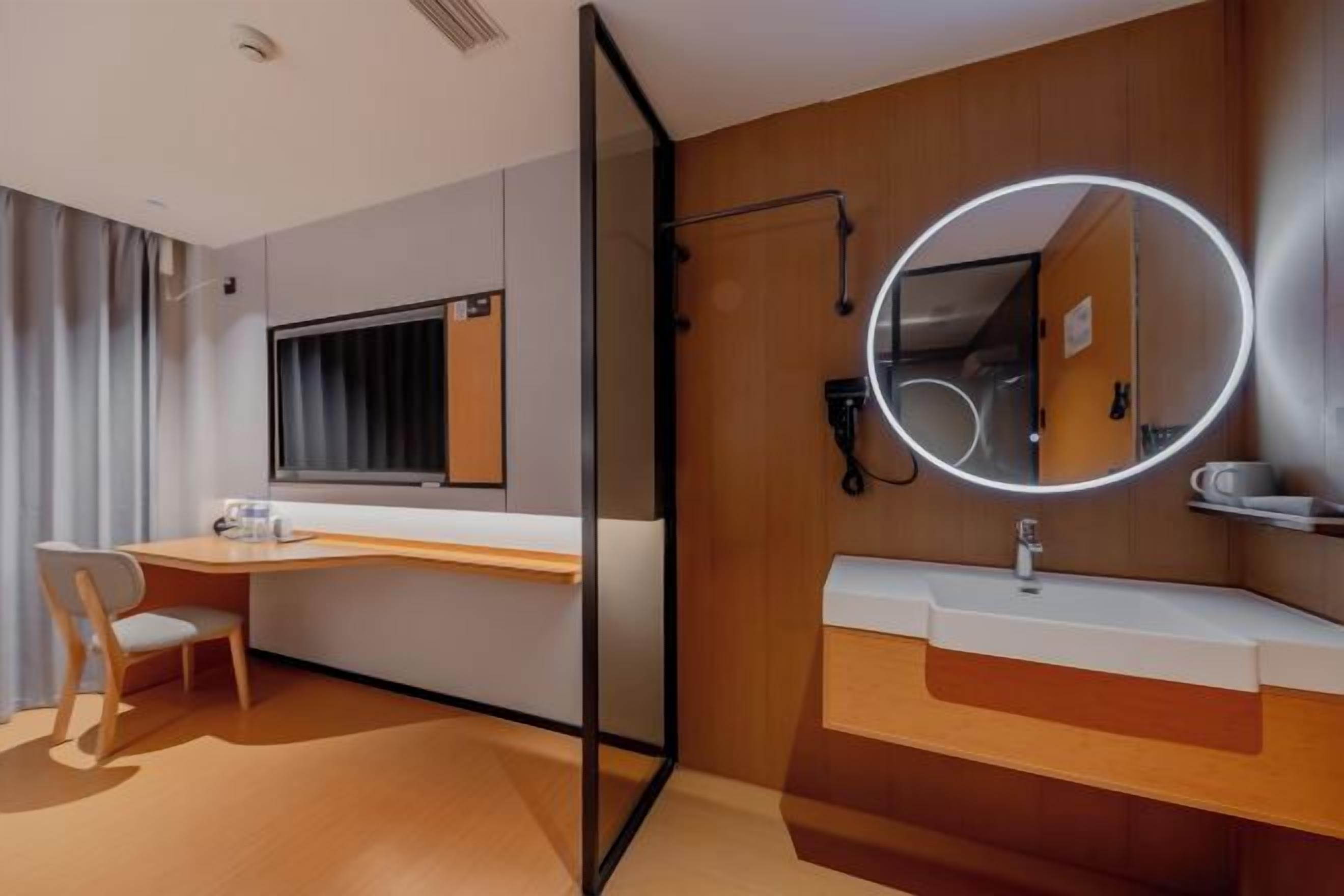 Una habitación de hotel moderna con un tocador de madera, un espejo redondo con luces LED, un escritorio y una silla.