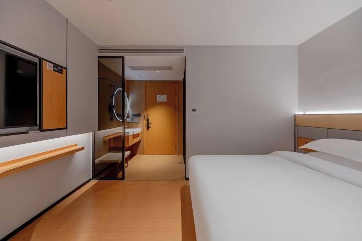 A modern hotel room with a bed, a TV, a mirror, and a bathroom with a shower.