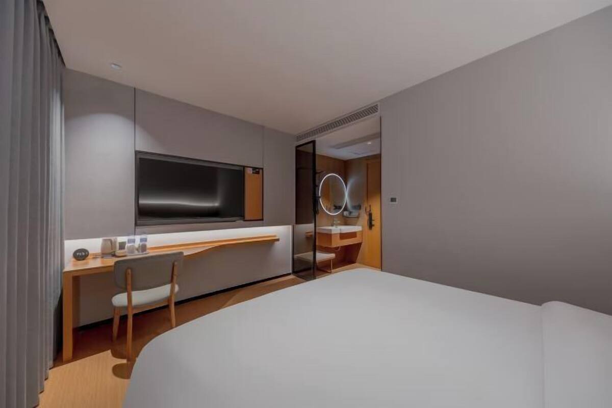 A modern hotel room with a large bed, a desk with a chair, a television, a mirror with lights, and a bathroom with a sink and mirror.
