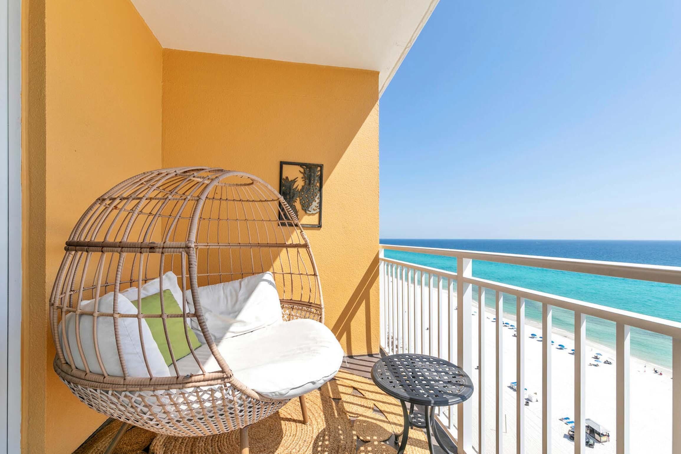 A balcony with a wicker chair, a small table, and a view of the beach.