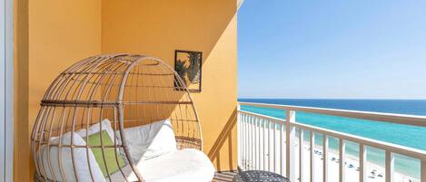 A balcony with a wicker chair, a small table, and a view of the beach.
