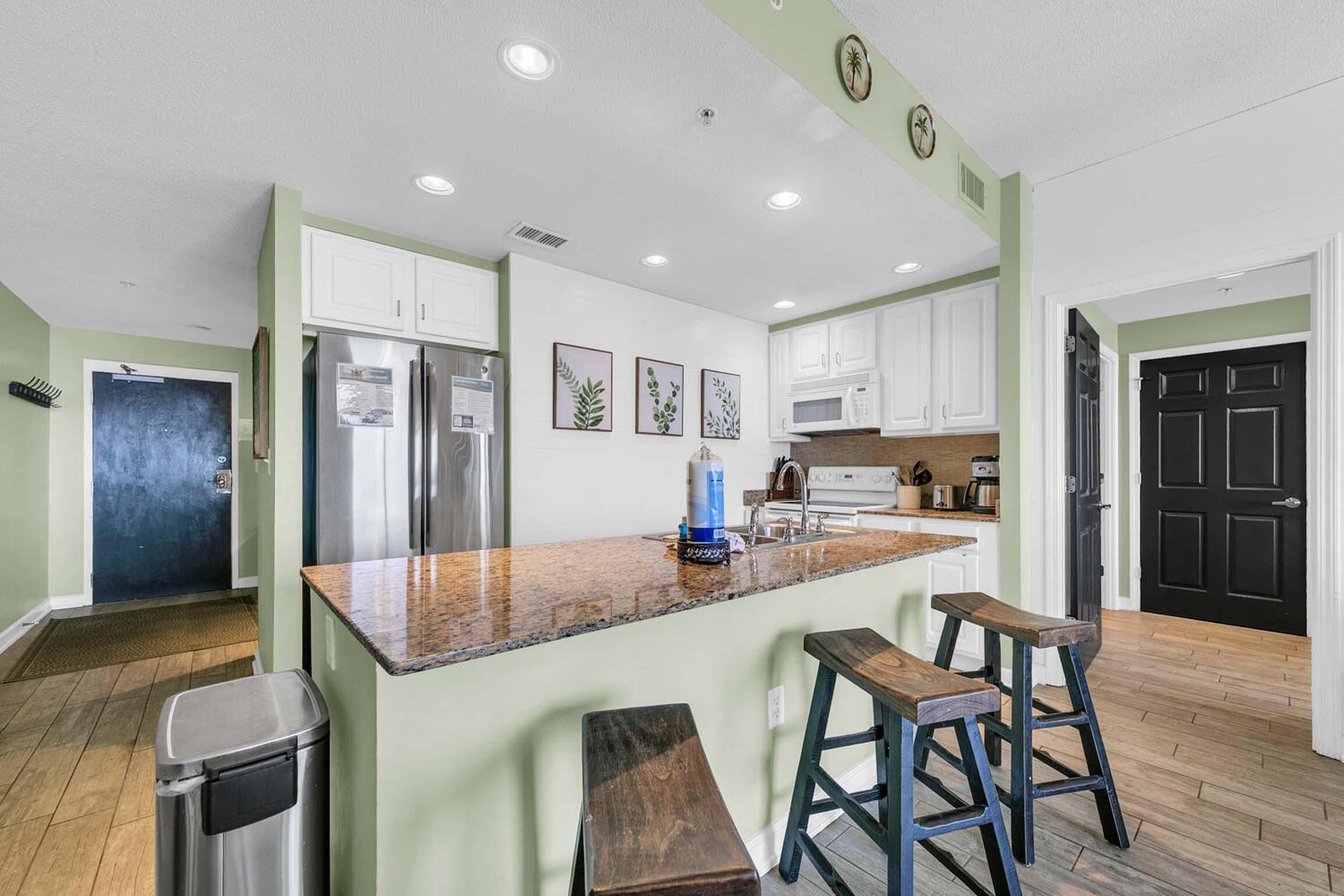A kitchen with white cabinets, a stainless steel refrigerator, a granite countertop, and wooden stools.