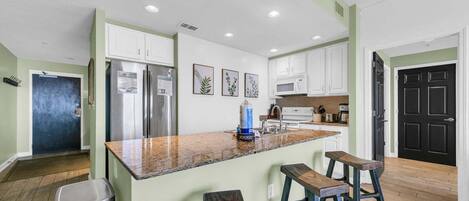 A kitchen with white cabinets, a stainless steel refrigerator, a granite countertop, and wooden stools.