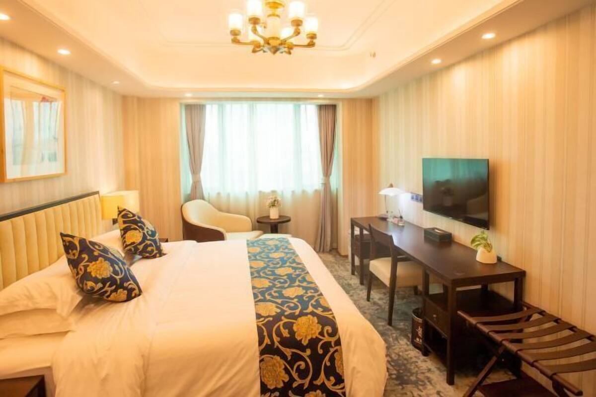 A hotel room with a bed, a desk, a chair, a TV, and a chandelier.