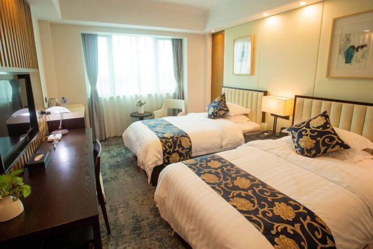 A hotel room with two beds, a desk with a television, a small table with a lamp, and a window with curtains.