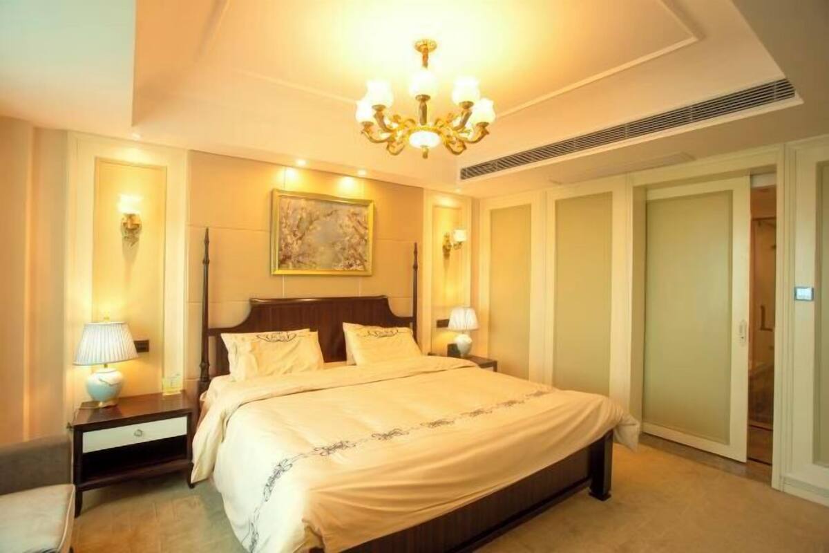 A hotel room with a large bed, a chandelier, a painting on the wall, and a door to the right.