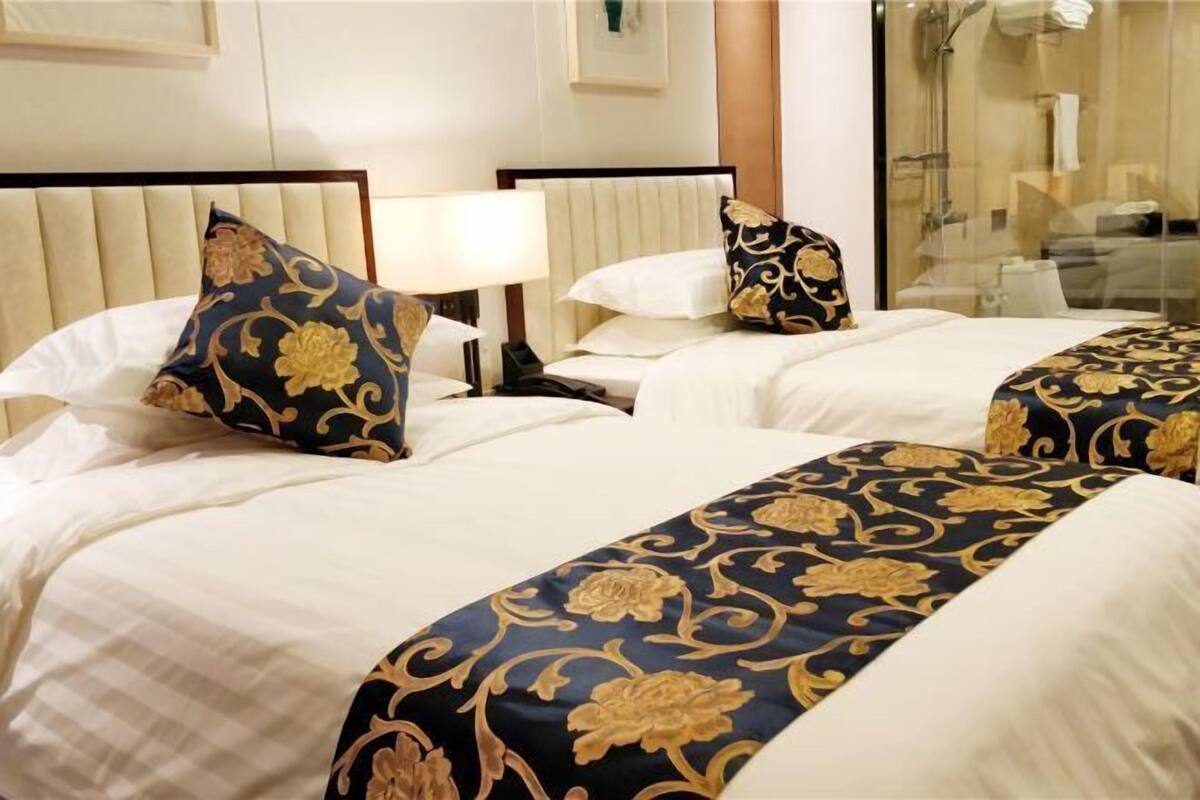 A hotel room with two beds, each with a white headboard and a blue and gold patterned bedspread.