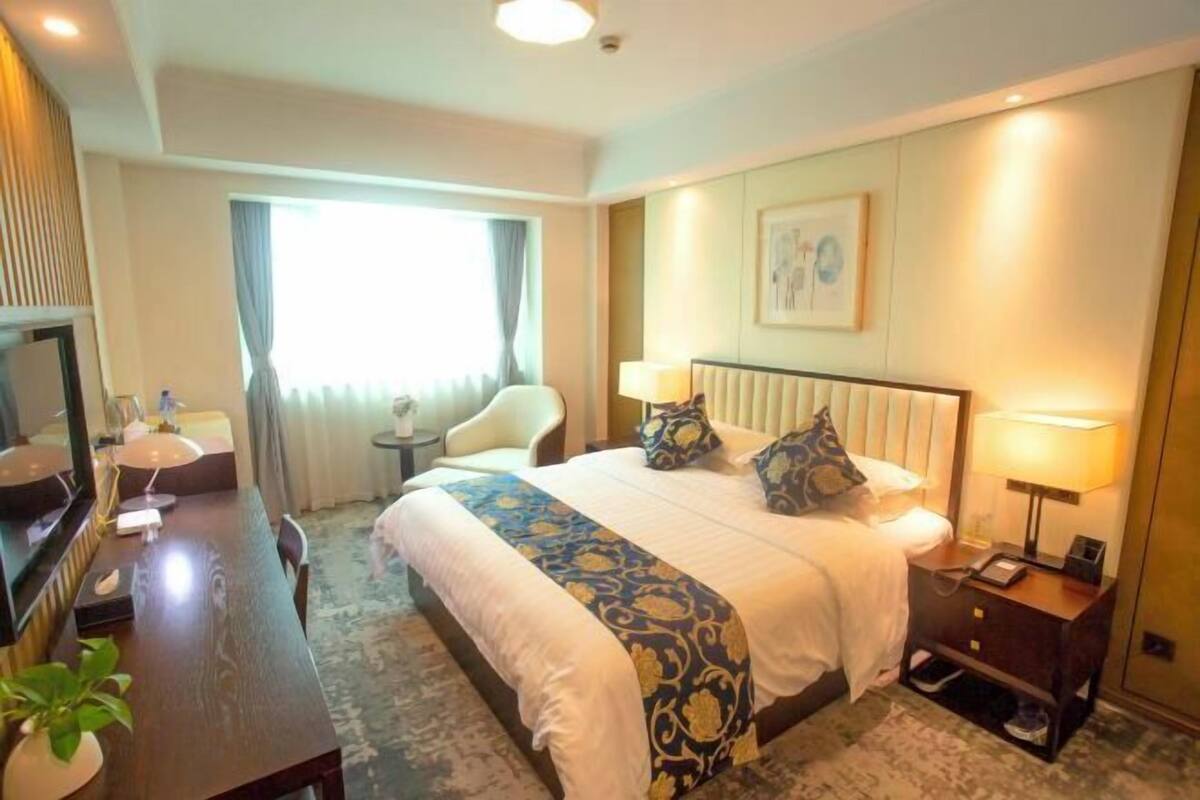 A hotel room with a bed, bedside table, lamp, chair, and a desk with a mirror and plant.
